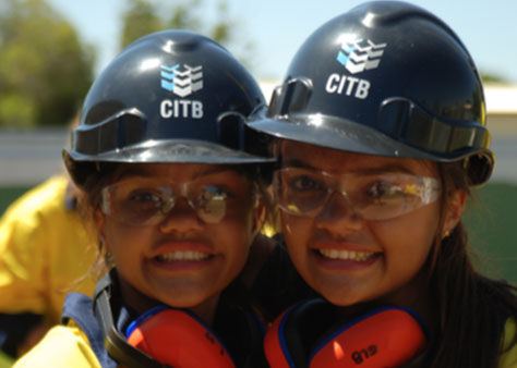 Image shows two girls wearing hard hats and smiling at the camera.